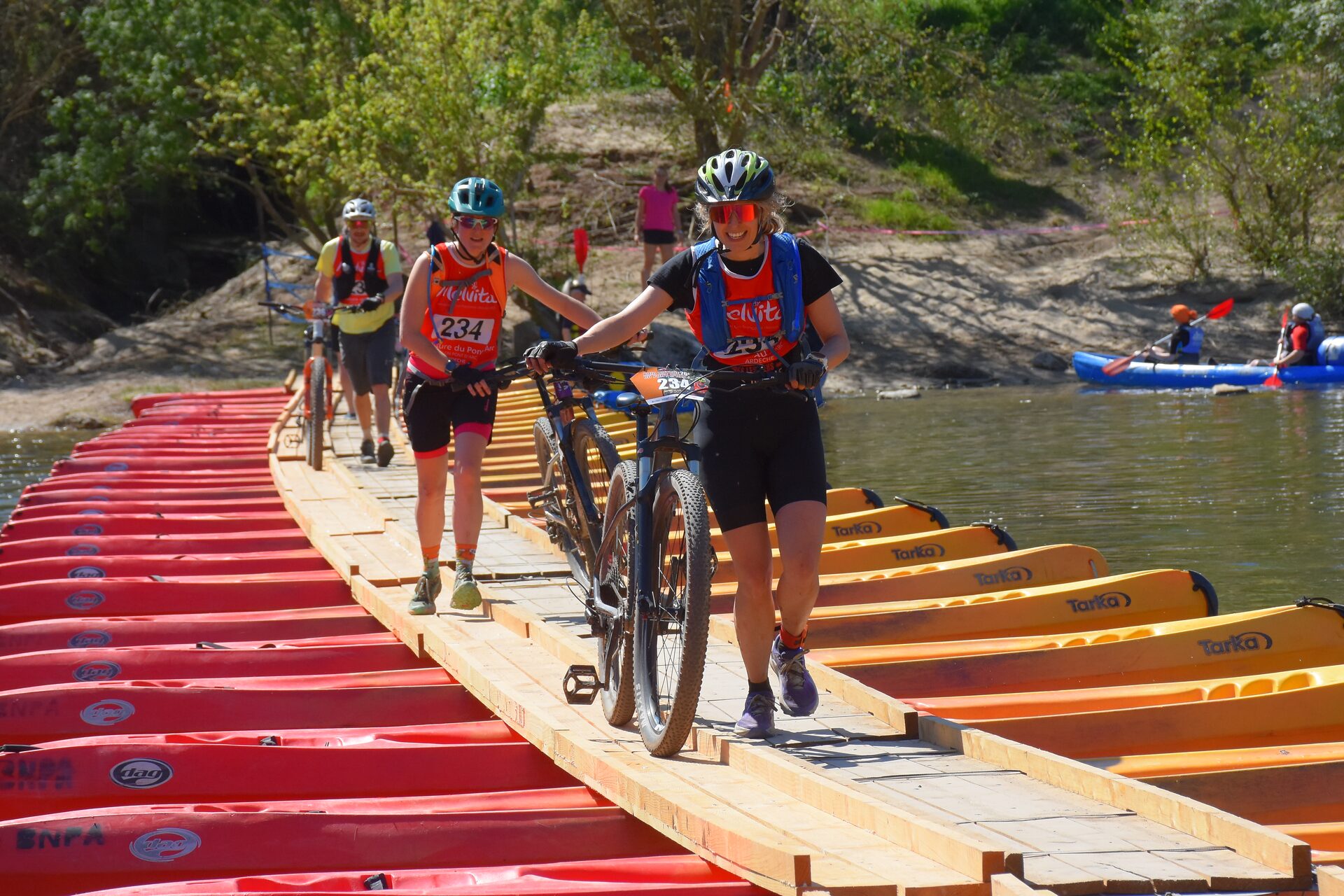 Femme souriante traversant un pont de bateaux à côté de son VTT lors du raid en équipe en Ardèche